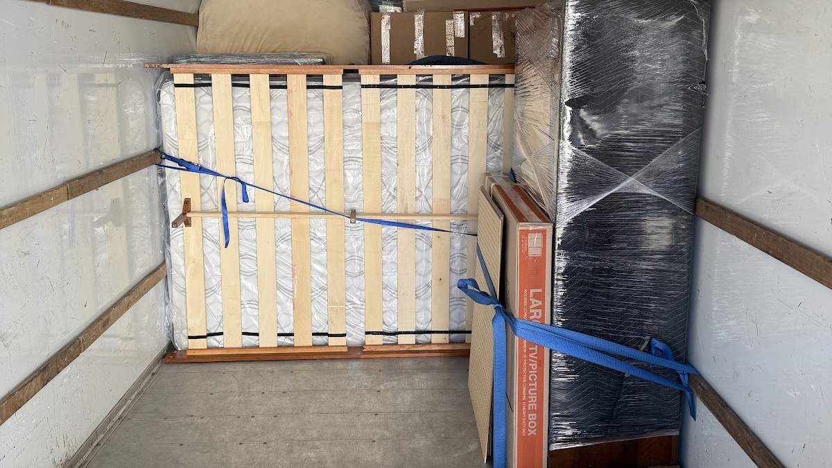 The inside of a moving truck mid-load: a bed frame and mattress wrapped in plastic, a furniture piece in a padded black wrap, and a TV box, all held against the wall with blue ratchet straps.