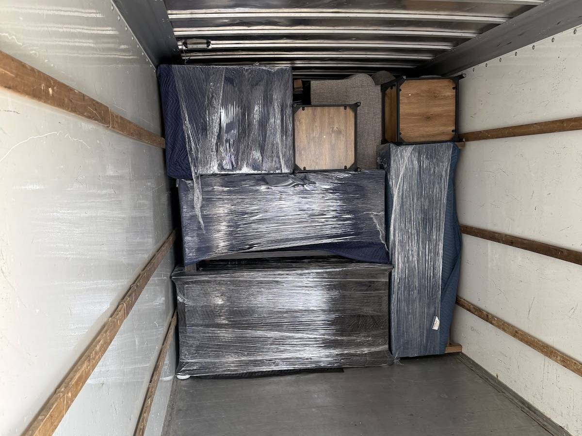 The back of a box truck loaded with furniture in blue padded blankets and black stretch wrap, with side tables stacked on top.